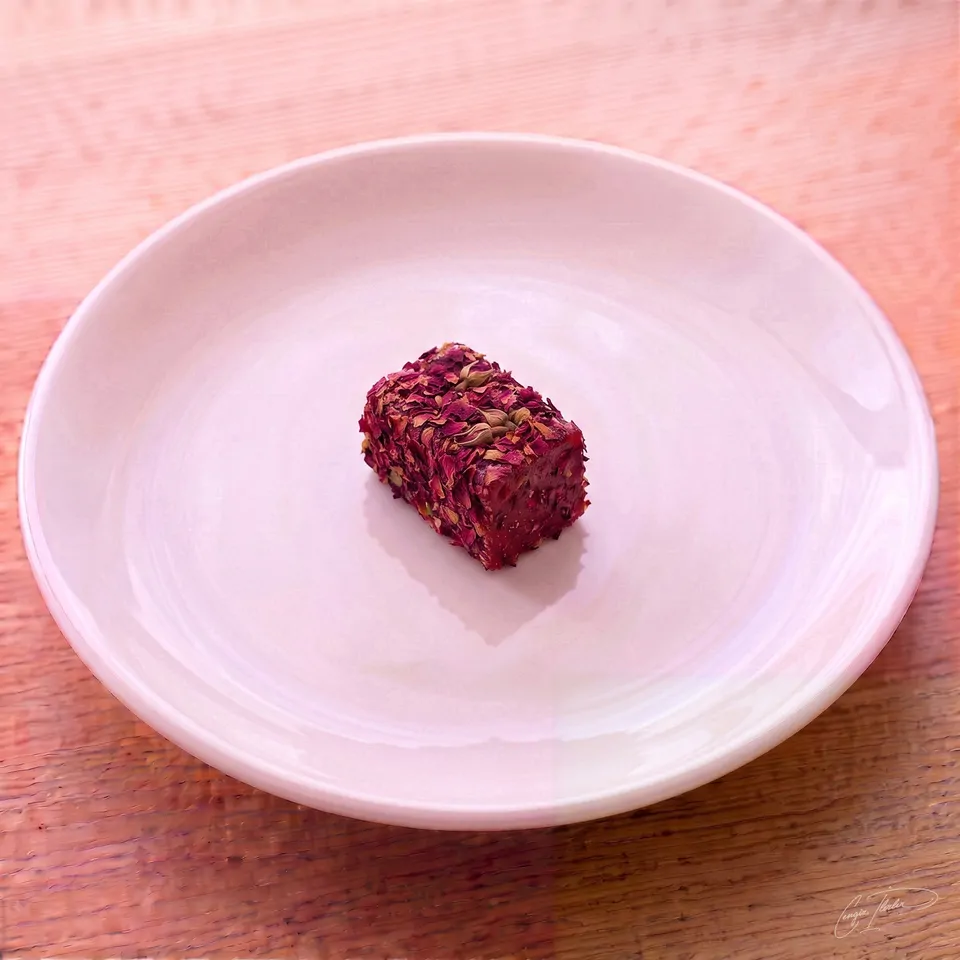 A single piece of lokum entirely coated in dried, dark pink rose petals, resting on a white plate.