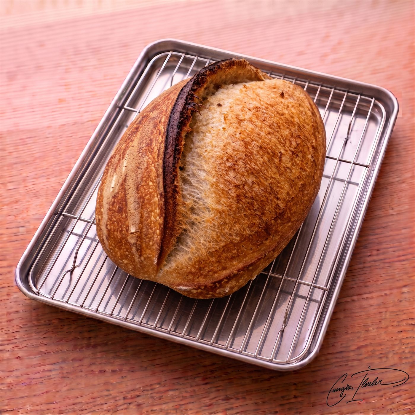 An oval-shaped loaf of crusty sourdough white bread featuring a prominent, deeply charred ear and scoring mark, resting on a wire cooling rack over a wooden surface.