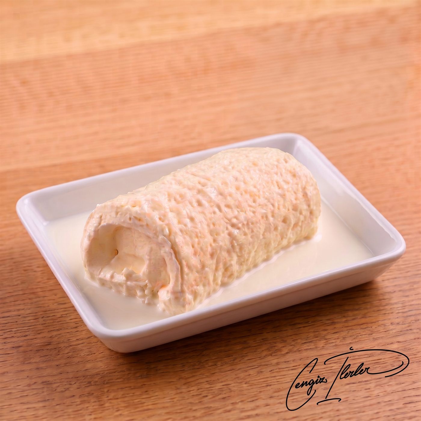 A rolled log of thick, creamy pale-yellow kaymak — Turkish clotted cream — resting in a shallow pool of milk in a white rectangular dish, on a wooden table.