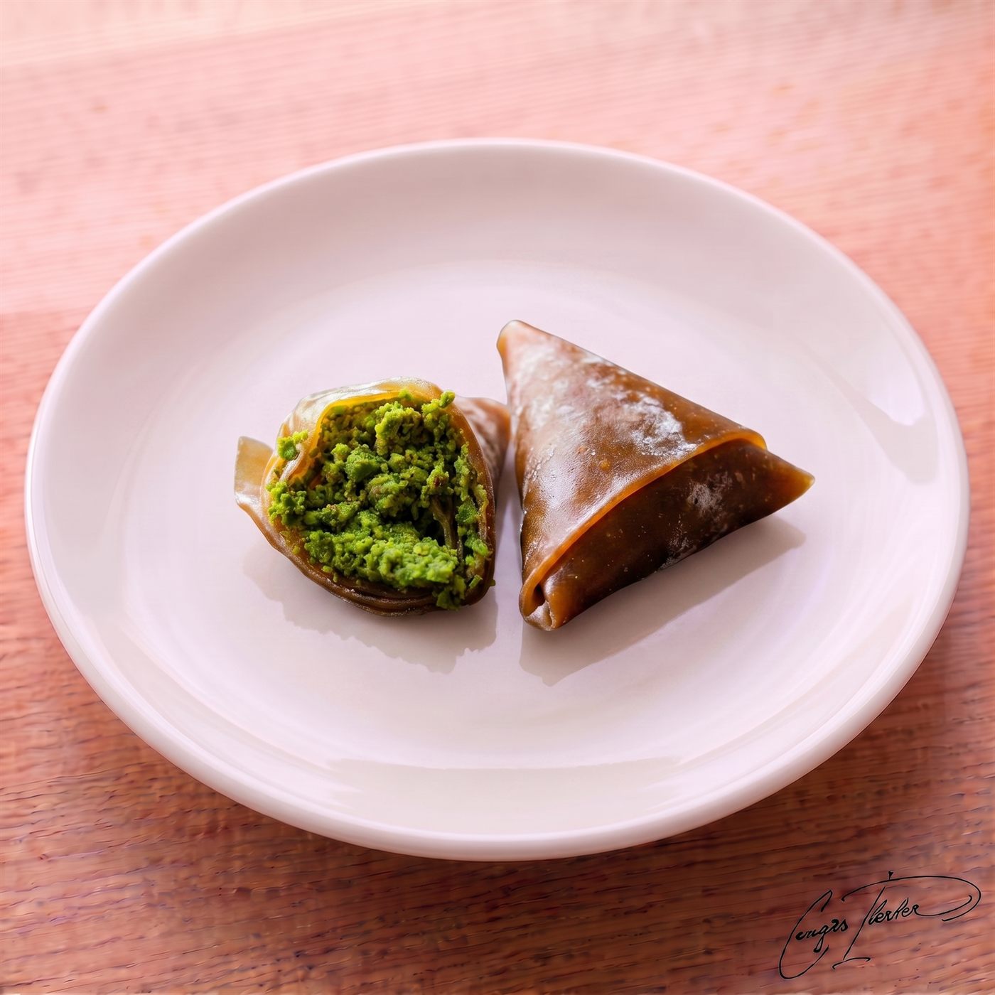 Two triangle-shaped muska (amulet) treats on a white plate, featuring a translucent, dark fruit-leather (pestil) wrapper folded over a dense, bright green pistachio filling.