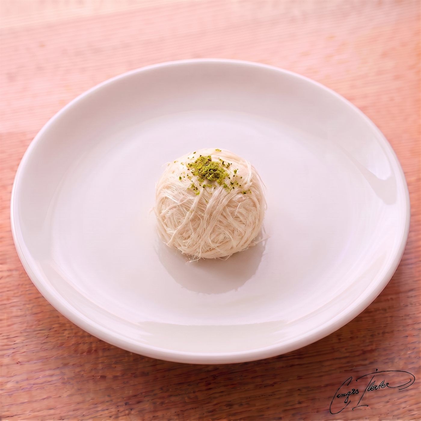 A single, delicate sphere of white, thread-like pişmaniye resting on a white plate, topped with a pinch of crushed green pistachios.