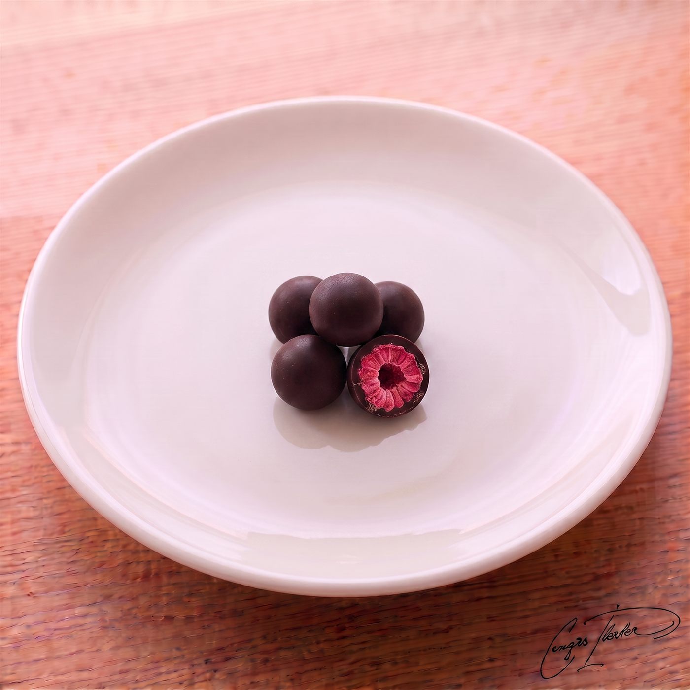 A small pile of dark chocolate-coated raspberry dragees on a white plate, with one piece cut open to reveal a bright pink, textured raspberry center.