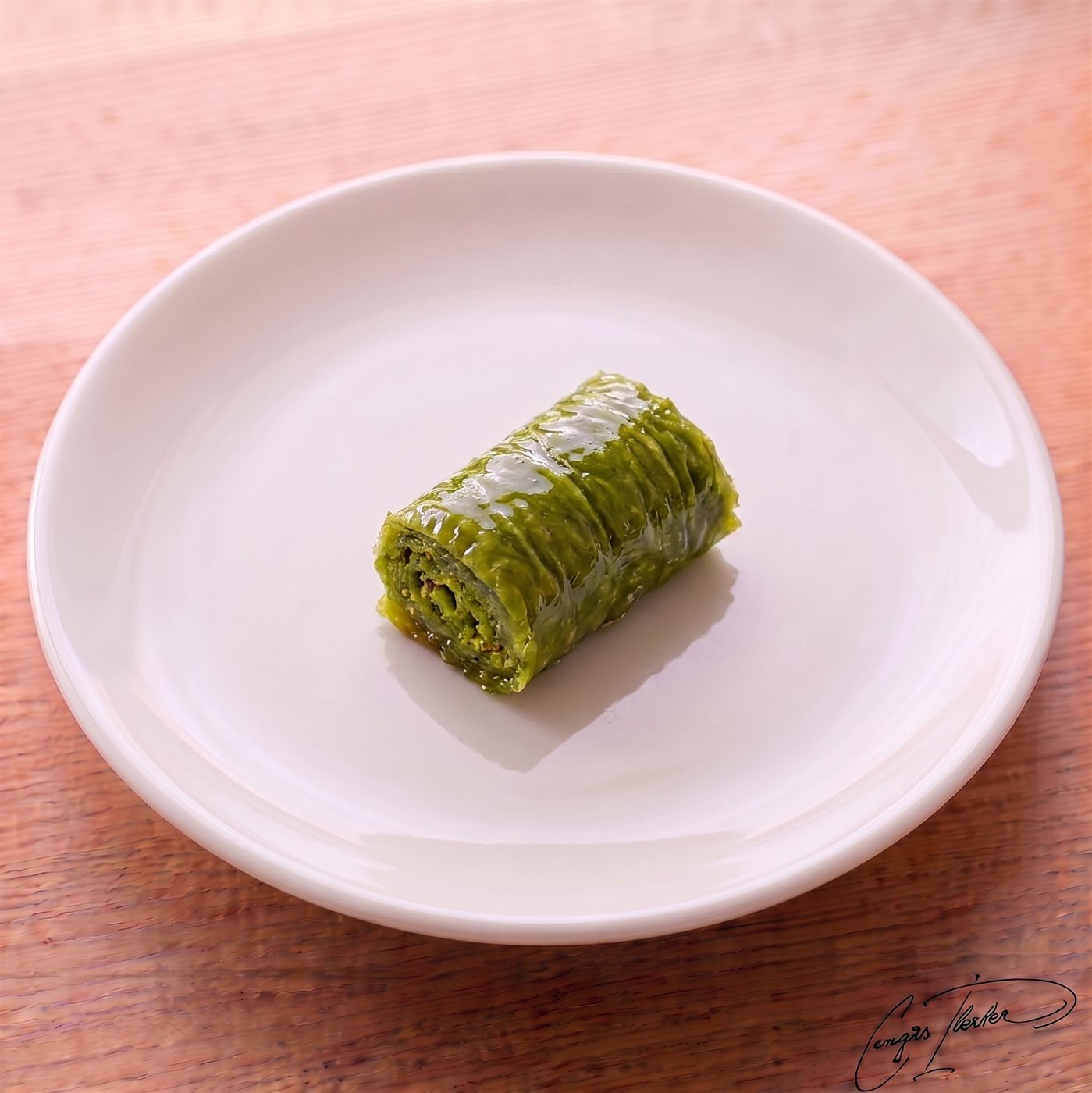 A single piece of fıstıklı dolama — a vivid green, tightly rolled Turkish dessert made almost entirely of ground pistachios wrapped in a single layer of phyllo dough and soaked in syrup — on a white plate.