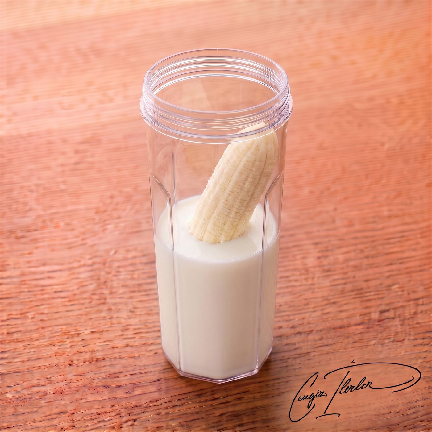 A clear blender cup containing fresh milk and a half-peeled banana standing upright inside it, resting on a wooden table.