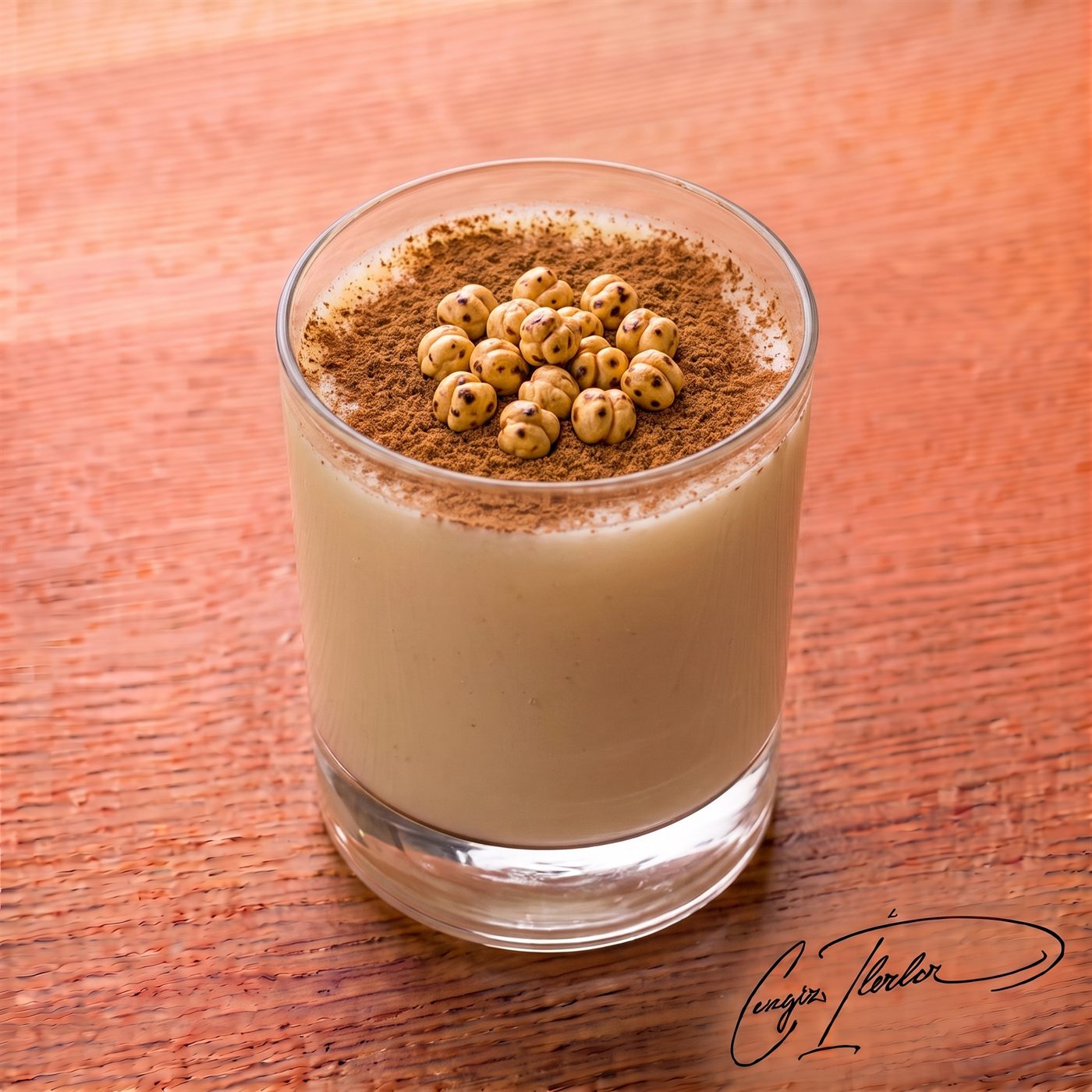 A tall glass of thick, pale-tan boza — the fermented millet drink — topped with cinnamon and roasted chickpeas, on a wooden table.