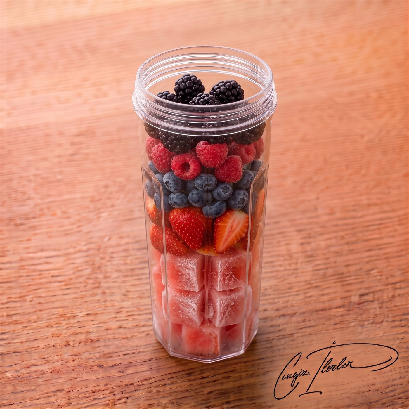 A clear tumbler filled with distinct layers of frozen watermelon cubes, sliced strawberries, blueberries, raspberries, and blackberries, ready to be blended, sitting on a wooden table.