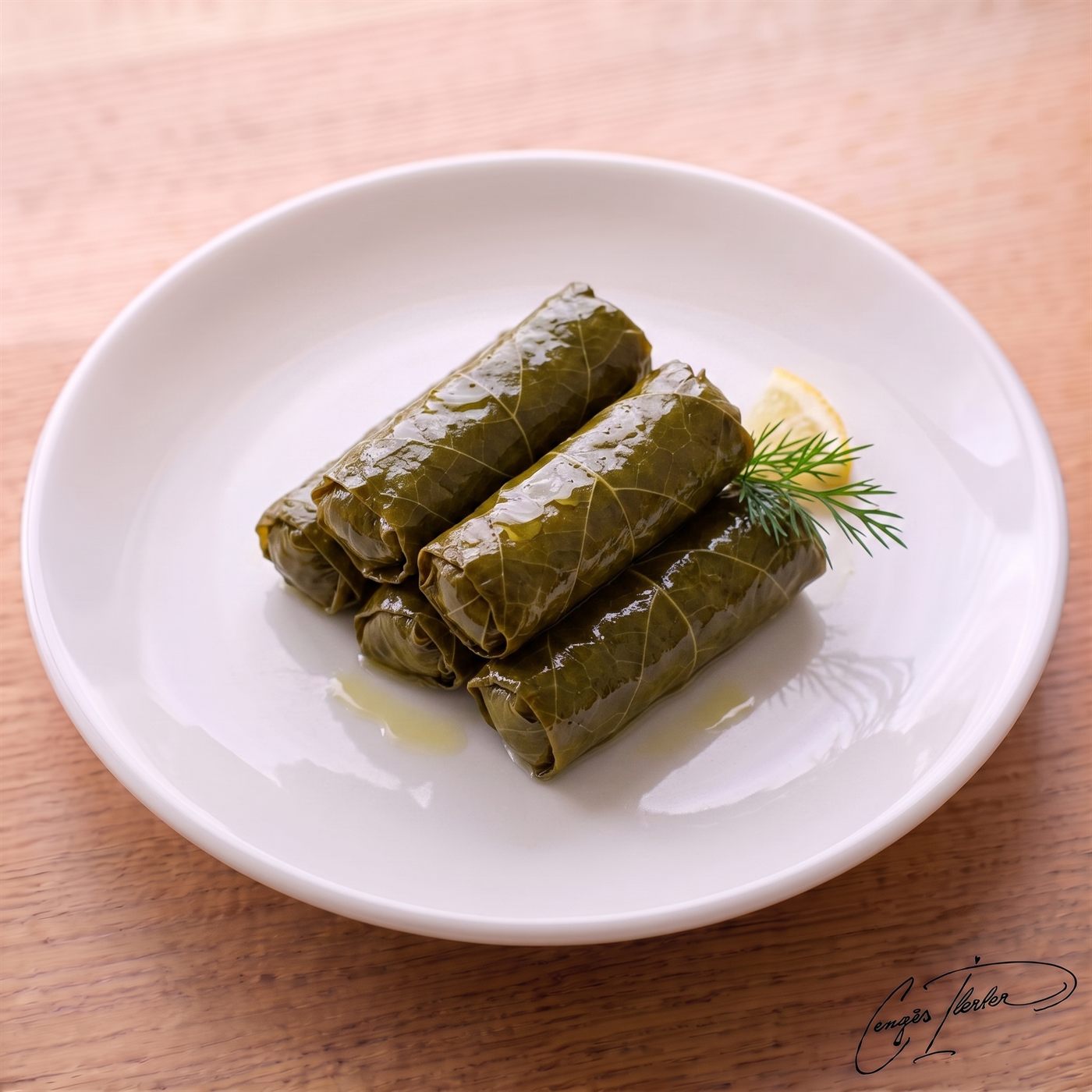 A neatly stacked pile of yaprak sarma (stuffed vine leaves) on a white plate, glistening with olive oil and garnished with a lemon slice and a sprig of fresh dill.