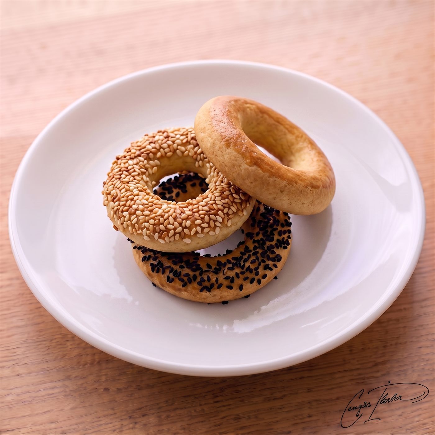 A stack of three small kandil simidi rings on a white plate — one plain, one coated in sesame seeds, and one coated in black nigella seeds.