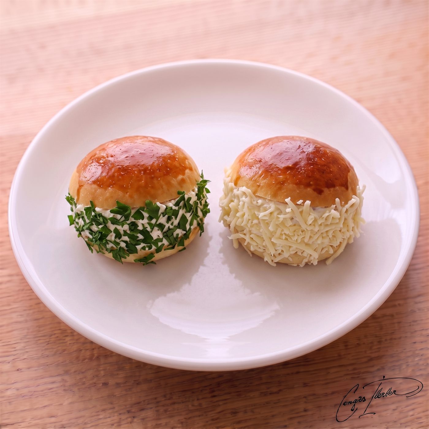 A white plate holding two mini Turkish 'bearded sandwiches' — small, shiny, golden-brown buns filled with cream cheese. The exposed filling of one is heavily coated in chopped fresh green parsley, and the other in grated yellow kashkaval cheese, creating their signature 'beards'.