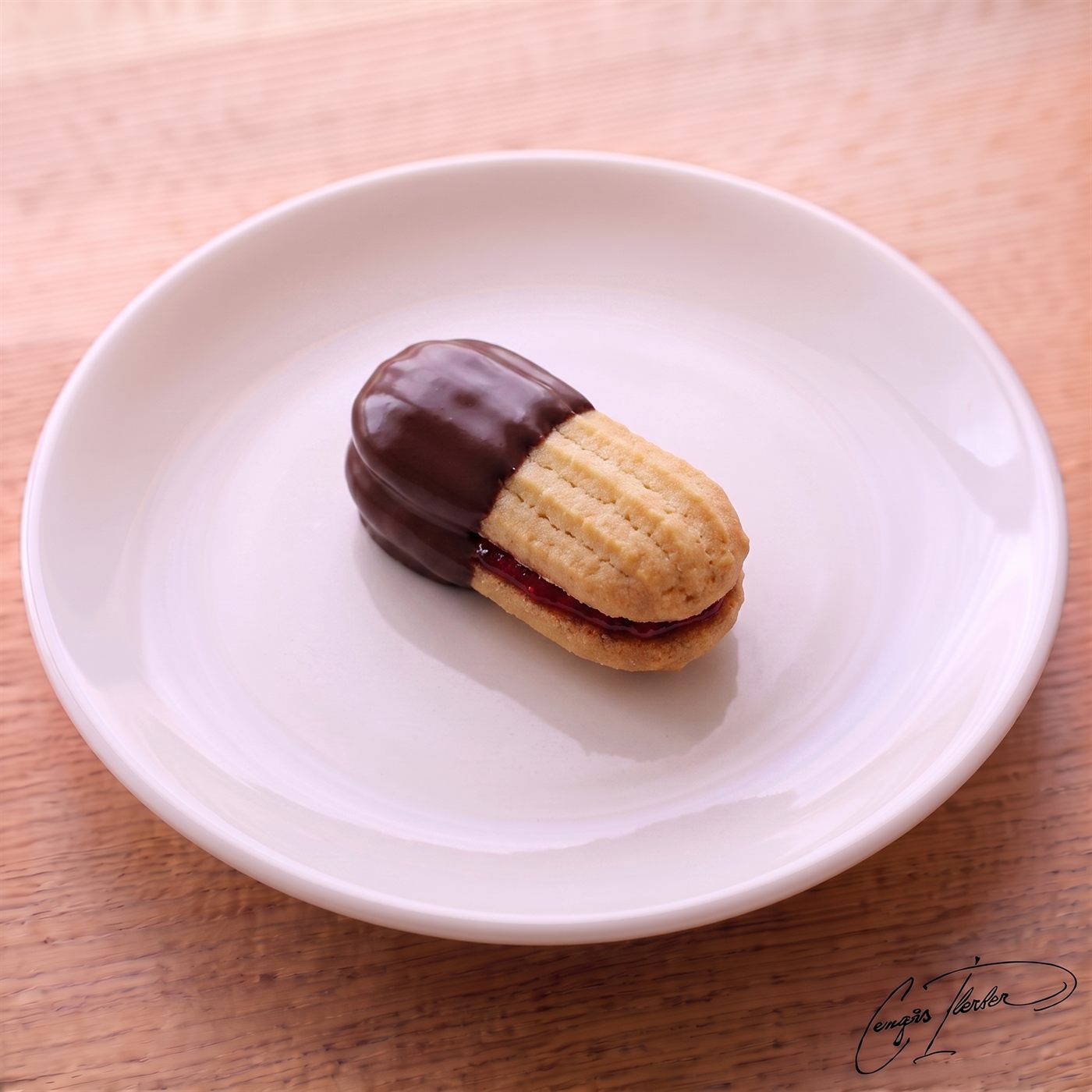A single tırtıl kurabiye on a white plate — two piped shortbread cookies sandwiched with red jam, with one end dipped in dark chocolate.
