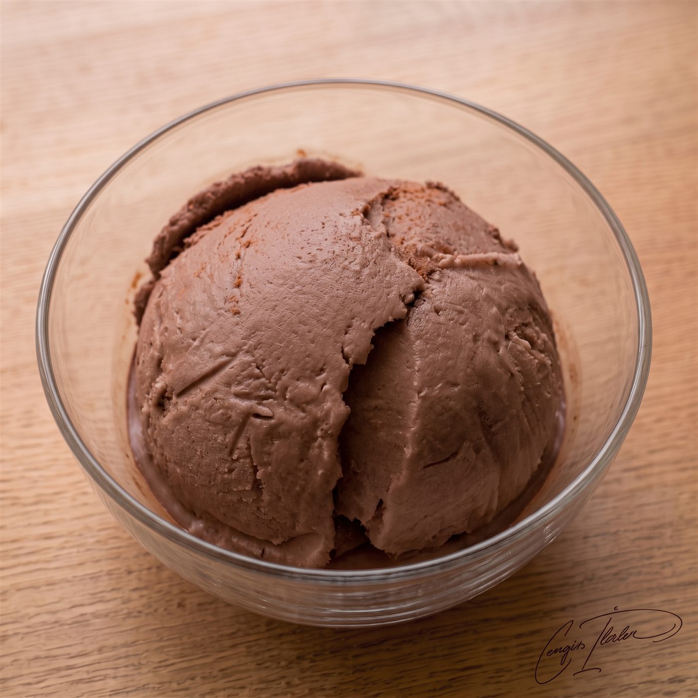 A single scoop of glossy chocolate ice cream in a clear glass bowl on a wooden table.