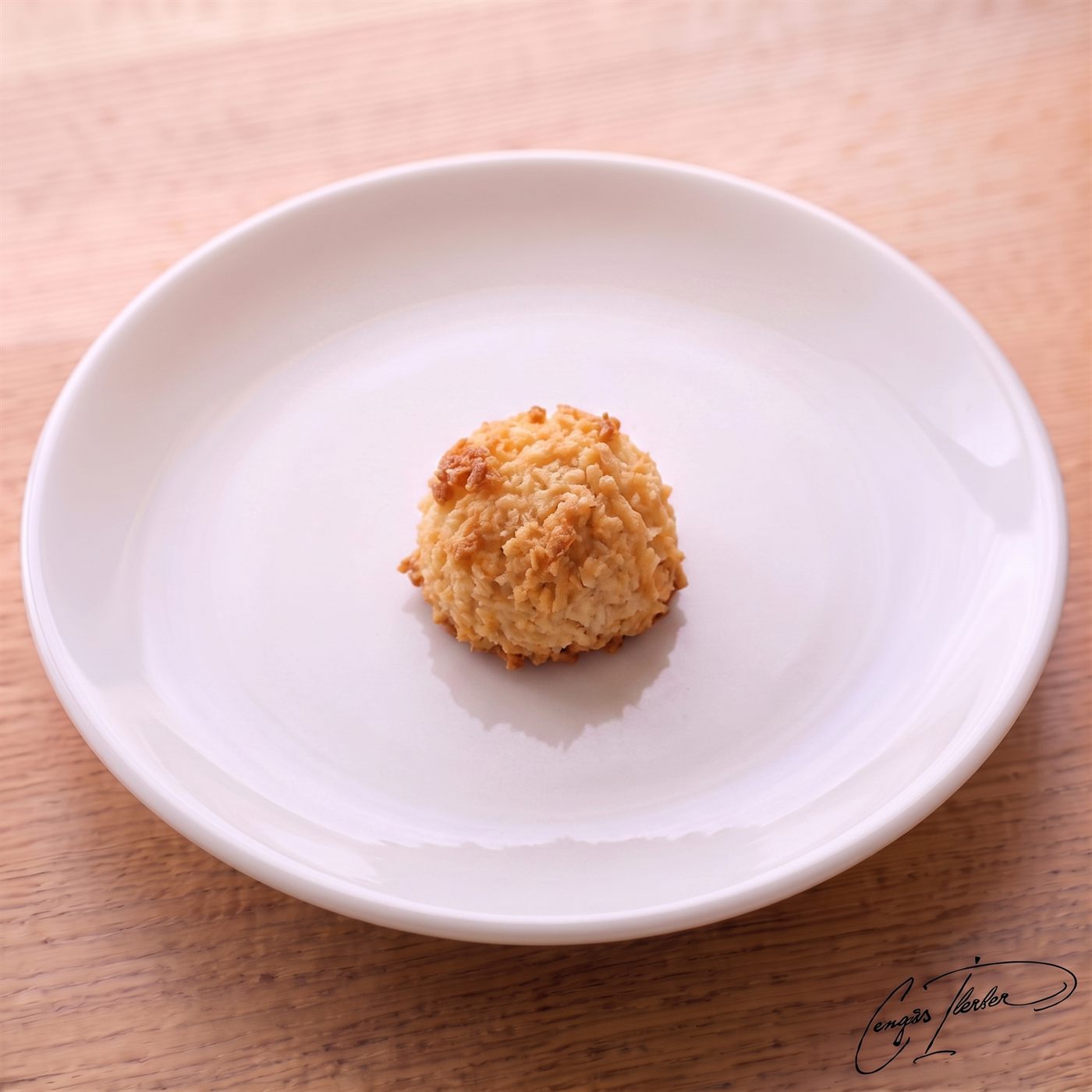 A single golden-brown coconut macaroon — a domed mound of baked shredded coconut — on a white plate on a wooden table.