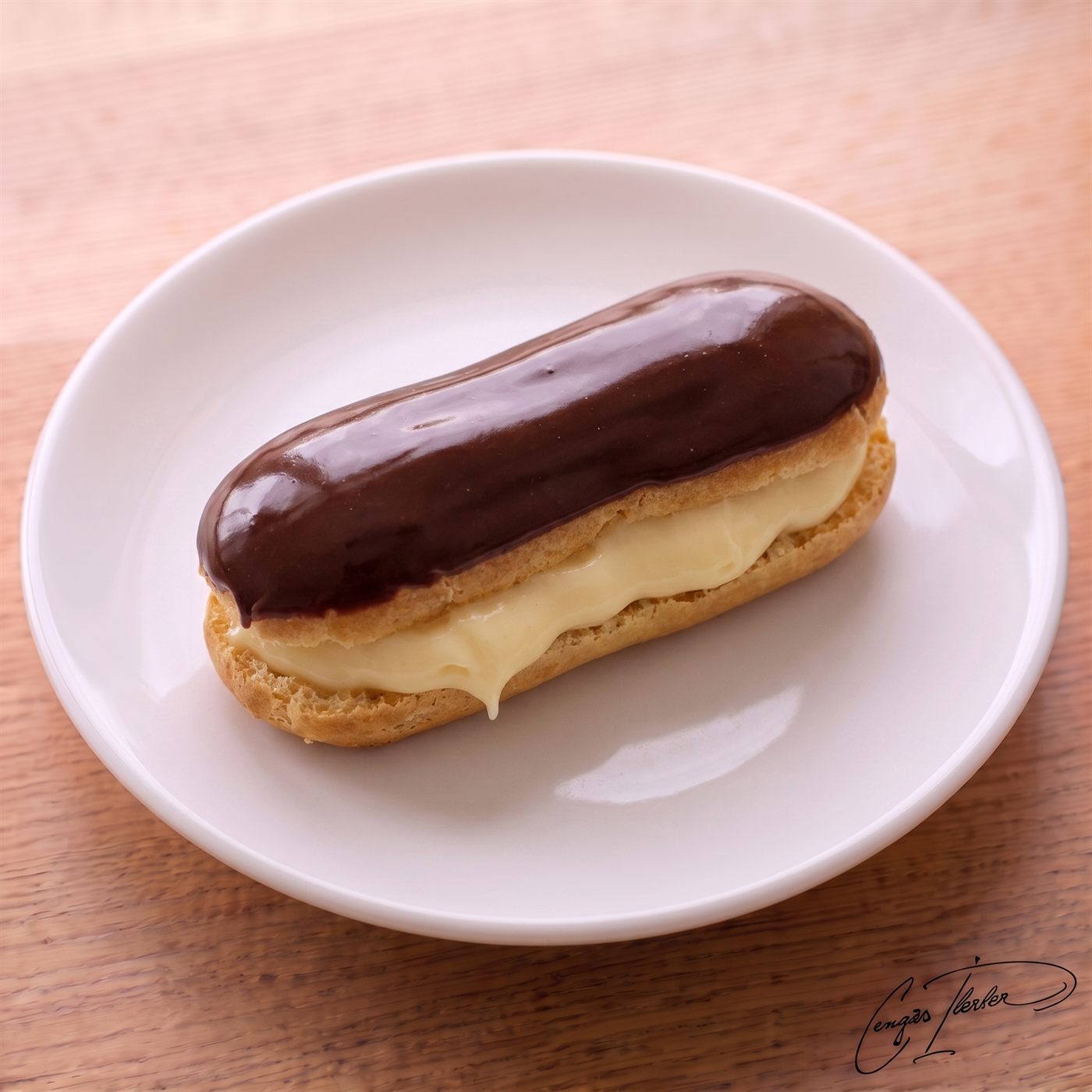 A row of chocolate-glazed éclairs filled with cream, on a white plate on a wooden table.