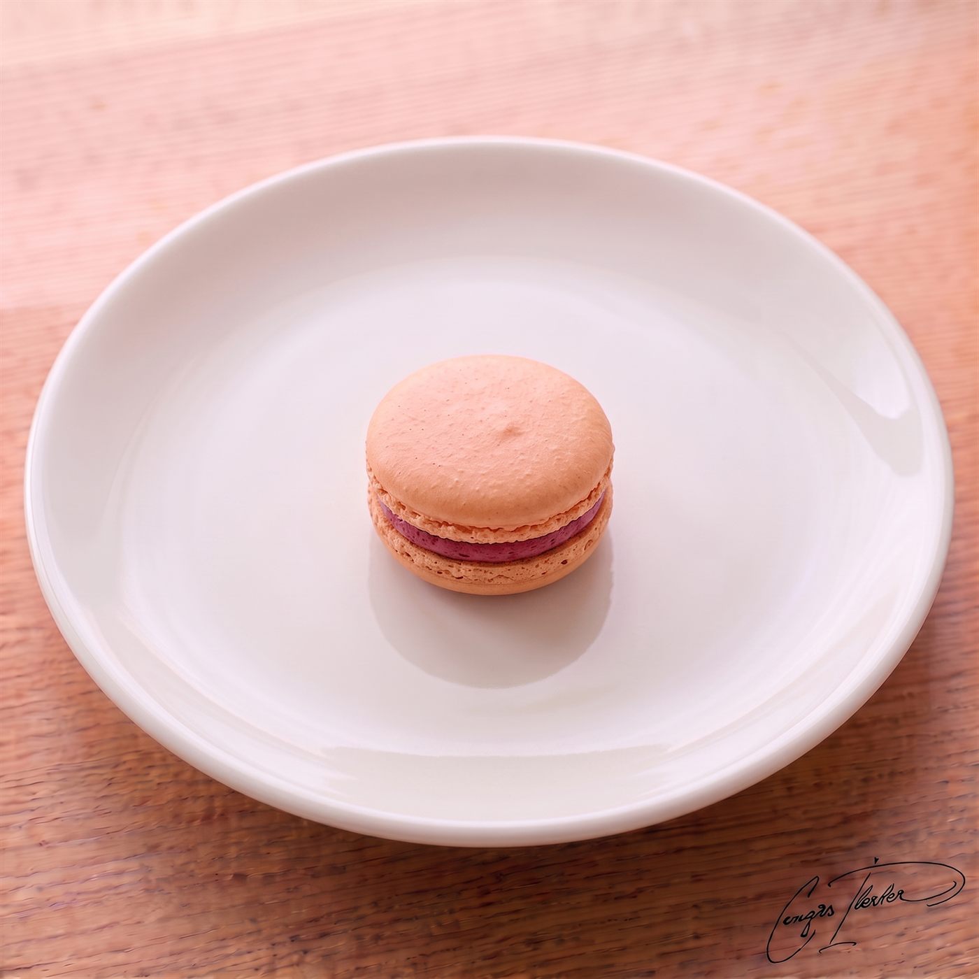 A single pale-orange French macaron — two smooth domed shells with ruffled feet — sandwiching a berry-pink filling, on a white plate on a wooden table.