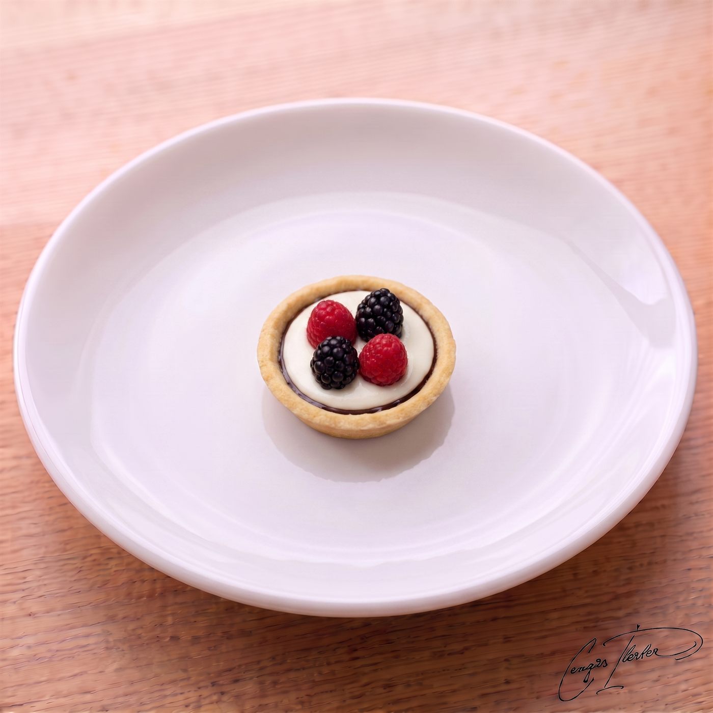 A small round tart with a golden shortcrust shell and a white-cream filling over dark chocolate, topped with two raspberries and two blackberries, on a white plate on a wooden table.