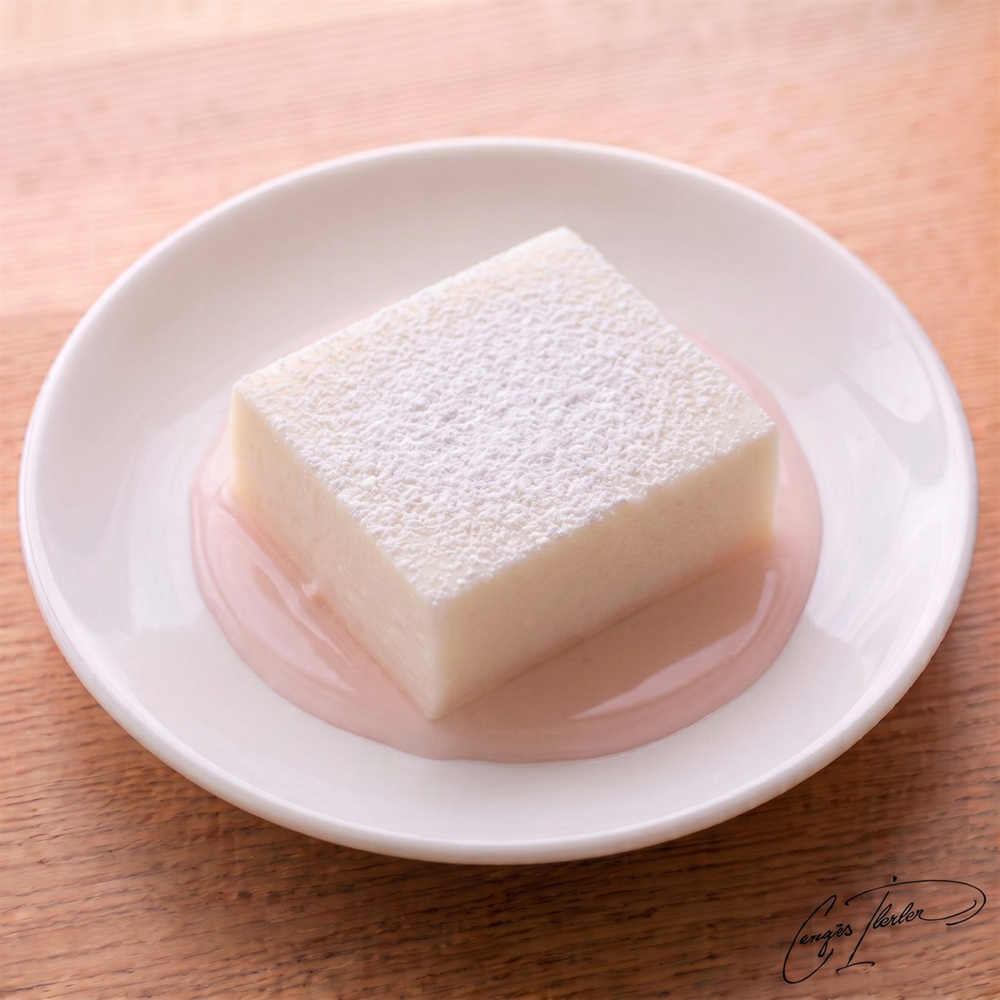 A perfect, translucent white square of su muhallebisi heavily dusted with powdered sugar, sitting in a pool of pale pink rosewater syrup on a white plate.