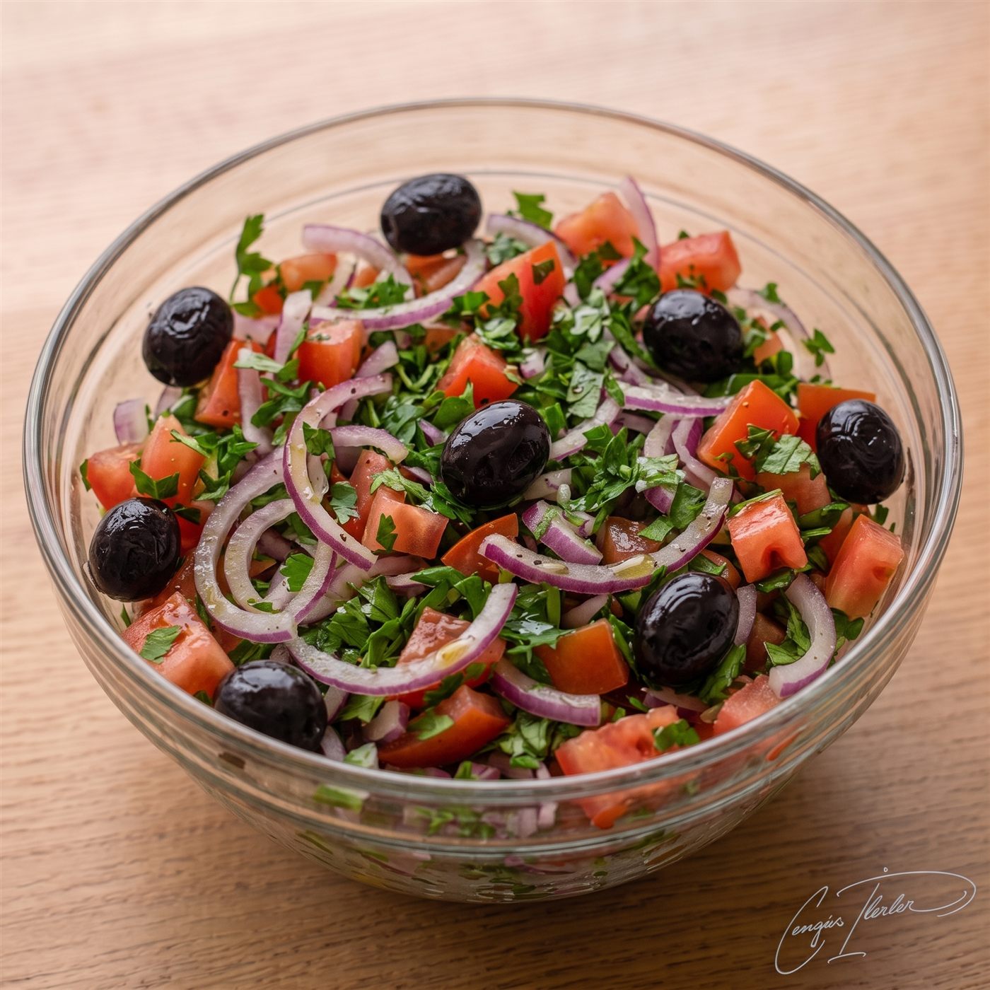 A glass bowl of çoban salatası — diced tomato, red onion half-moons, whole black olives, and plenty of parsley.