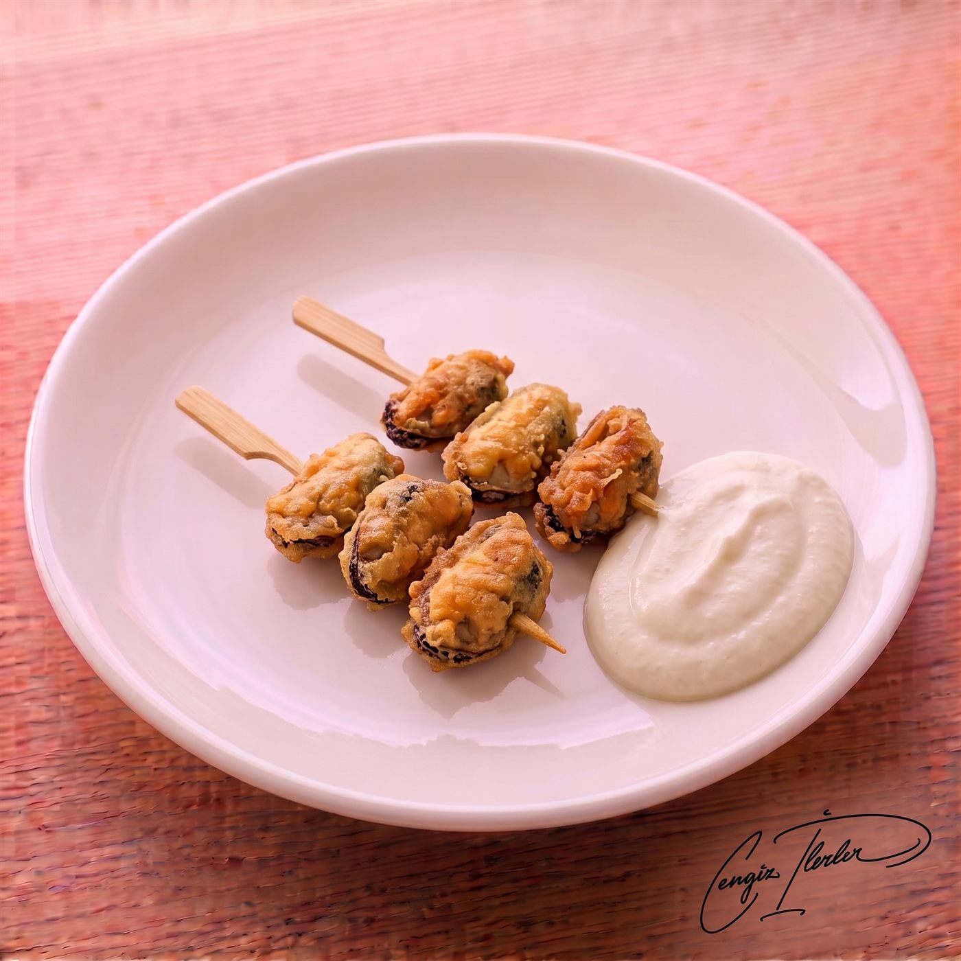 Two wooden skewers of golden, crispy midye tava (fried mussels) resting on a white plate beside a generous dollop of creamy tarator sauce.
